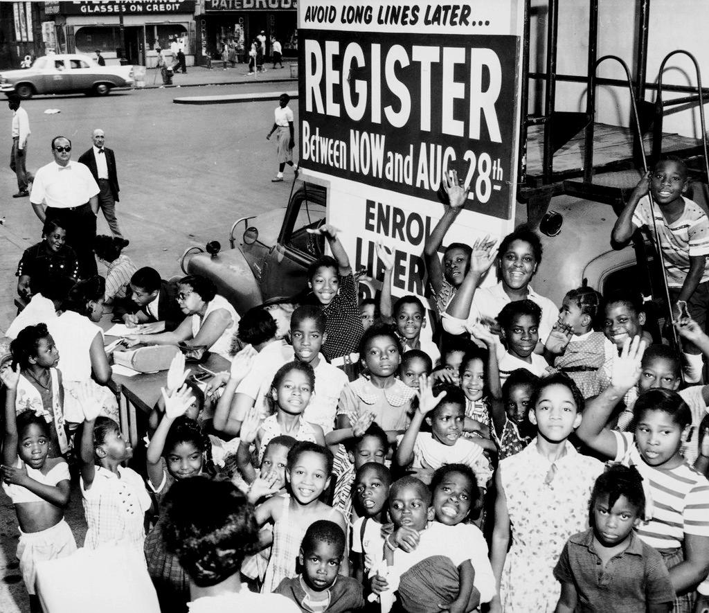 A group of African-American children gather around a sign and booth to register voters. Early 1960s.